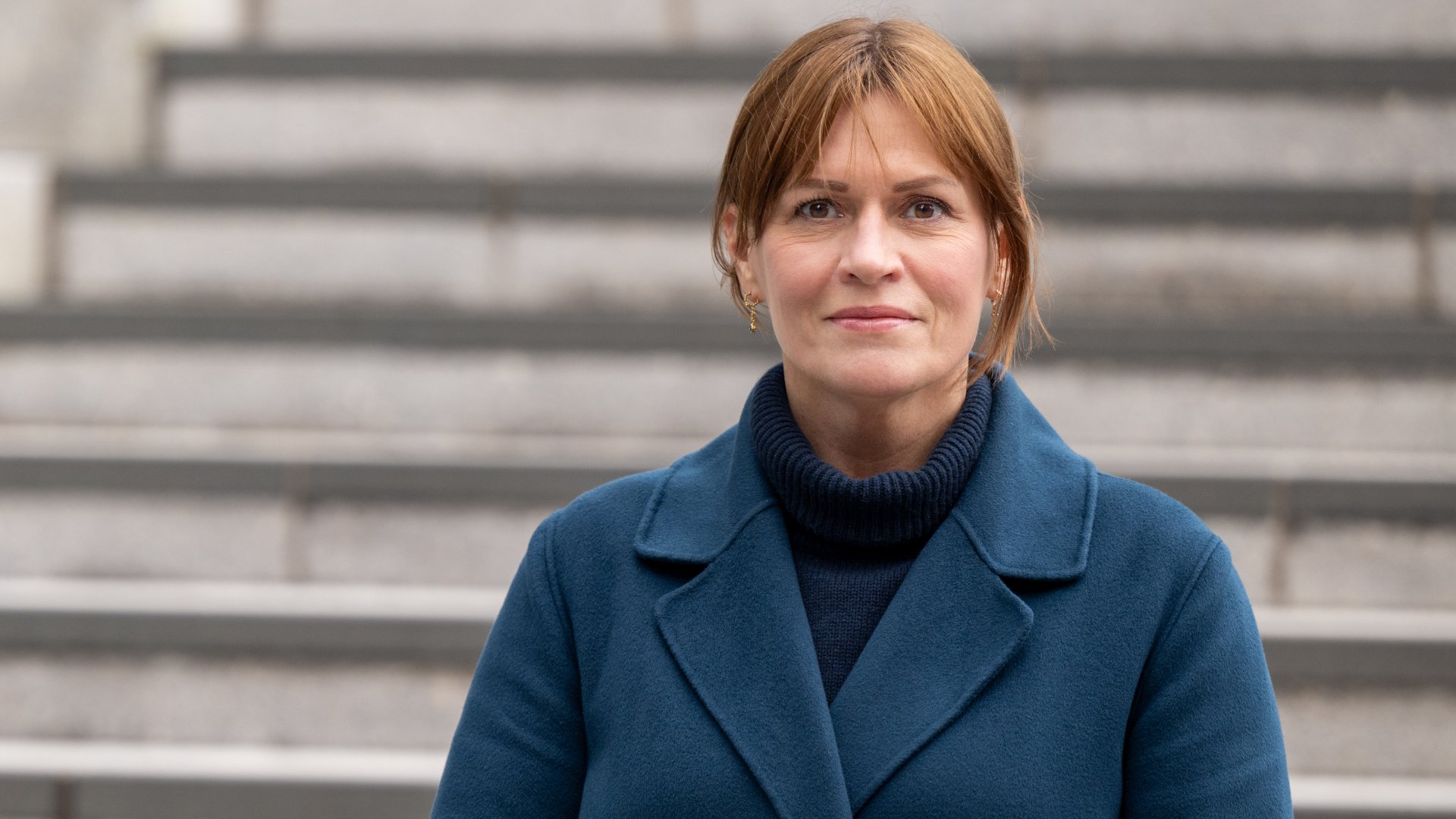 Kvinne med høyhalset genser, blå kåpe og brunt hår som ser inn i kamera