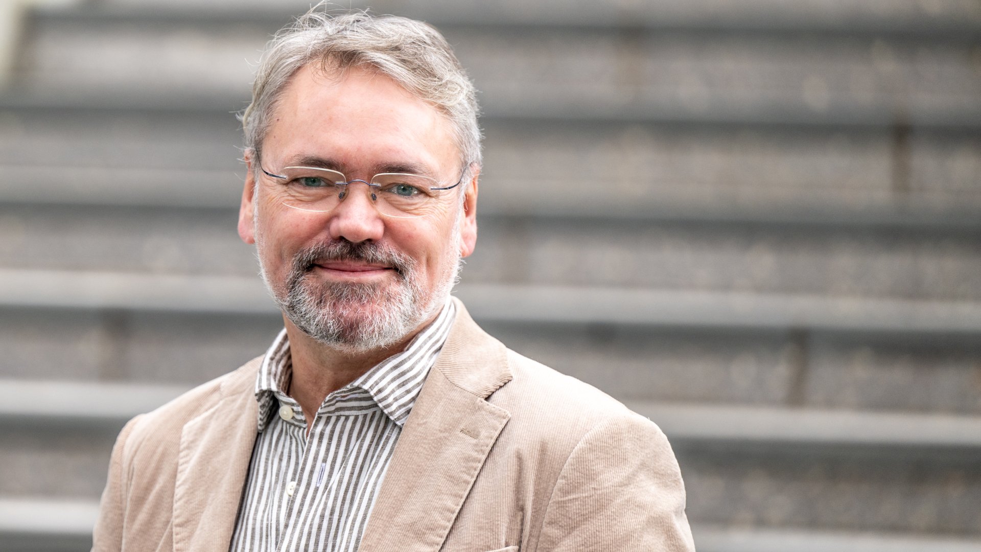 En mann med briller og grått skjegg, med lys blazer og stripete skjorte står foran grå trappetrinn.