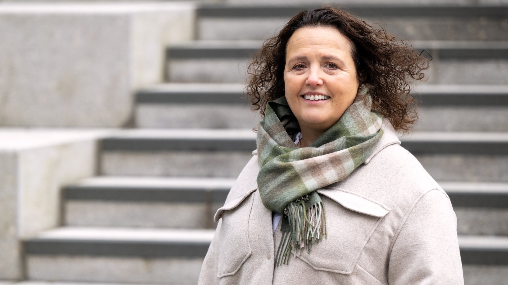 En kvinne med krøllete hår, lys frakk og rutete skjerf står foran grå trappetrinn.